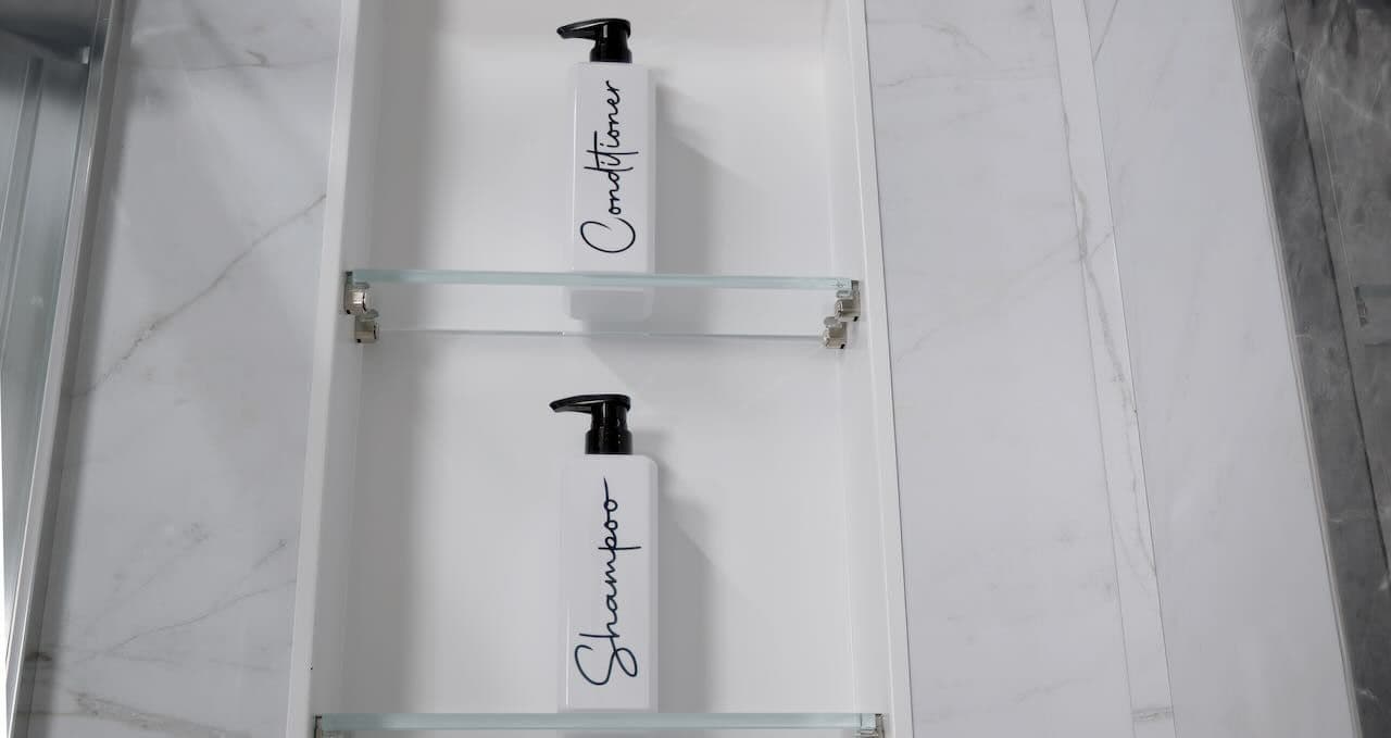 Two white bottles labeled "Shampoo" and "Conditioner" with black pumps are neatly placed on glass shelves in a sleek, modern shower.