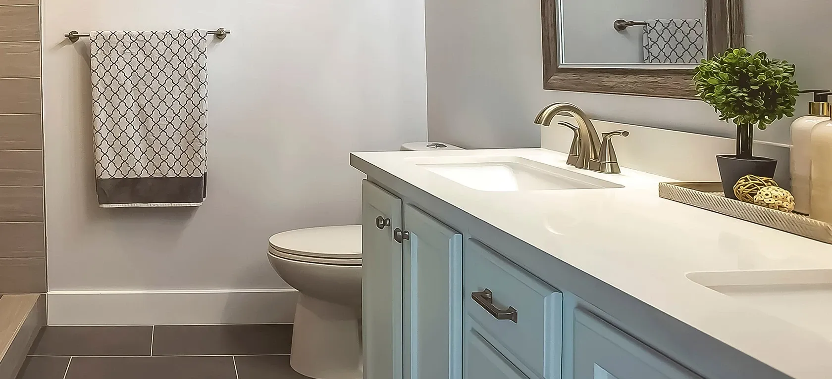 Bright, modern bathroom with a white vanity and a chrome faucet. A framed mirror and a plant sit on the counter. A patterned towel hangs nearby.