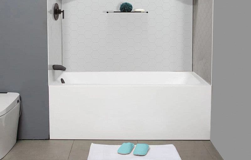 A white bathtub with hexagon tile wall, shelf, aqua slippers, and a white bath mat on the floor.