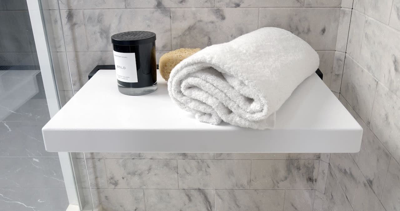 A white shower bench seat in a marble-tiled bathroom holds a rolled white towel, a beige sponge, and a black candle