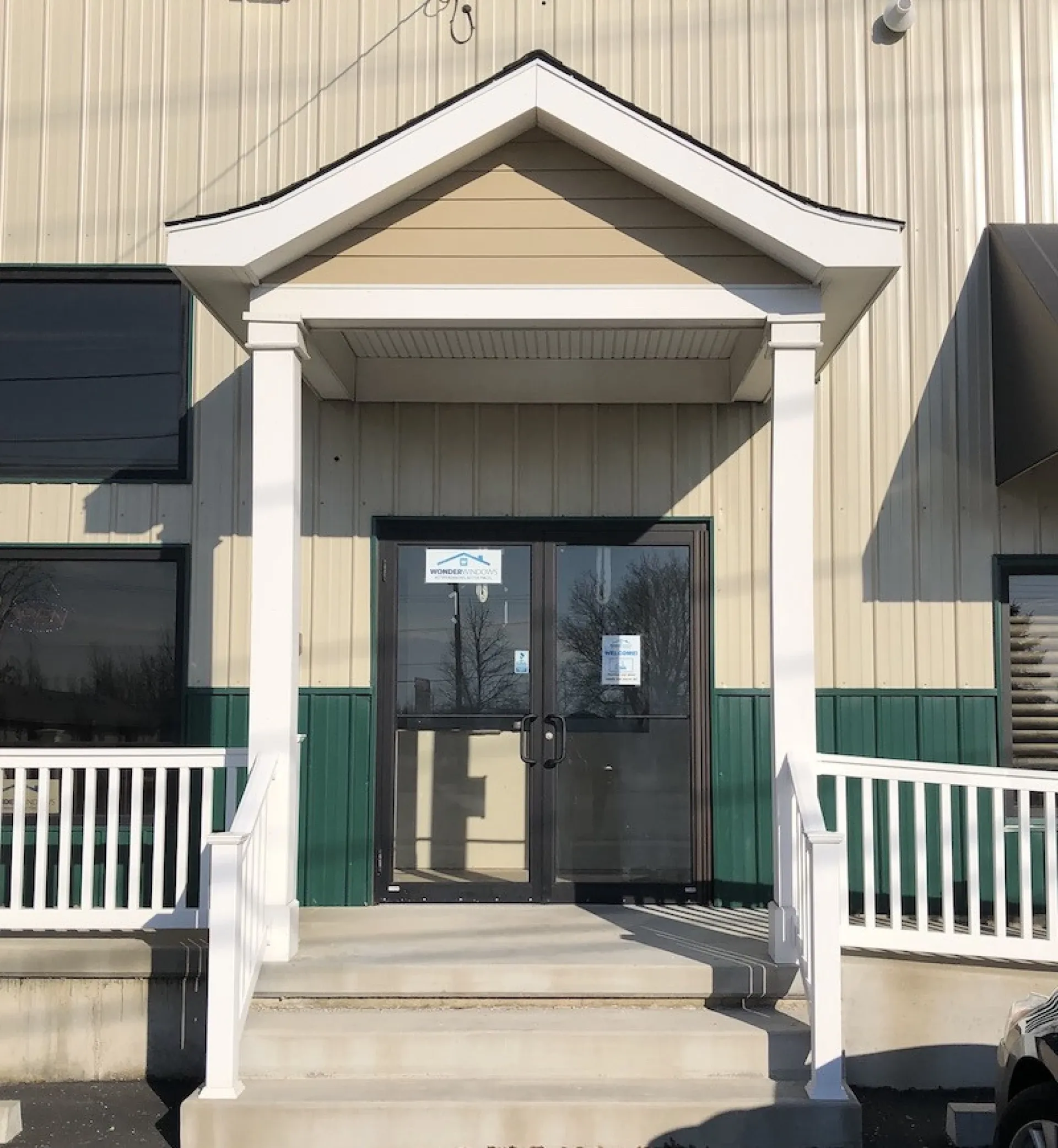 Entrance of a beige building with a gable roofed porch, white columns, and a green accent. Glass double doors reflect trees.