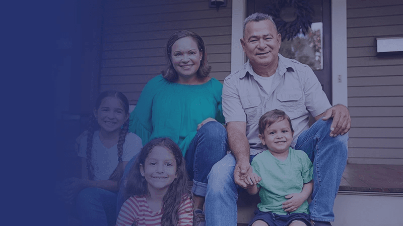 A smiling family of five sits on wooden steps in front of their home.