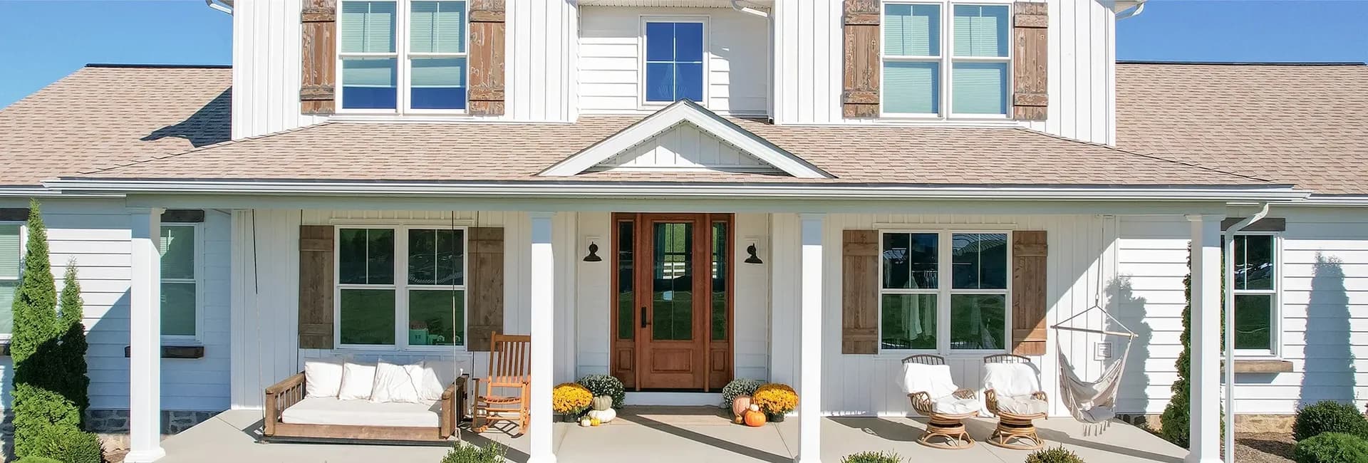 Front of a white, modern farmhouse with tan roof, wooden shutters, and a welcoming porch. Features cozy seating, potted plants, and a hammock chair.