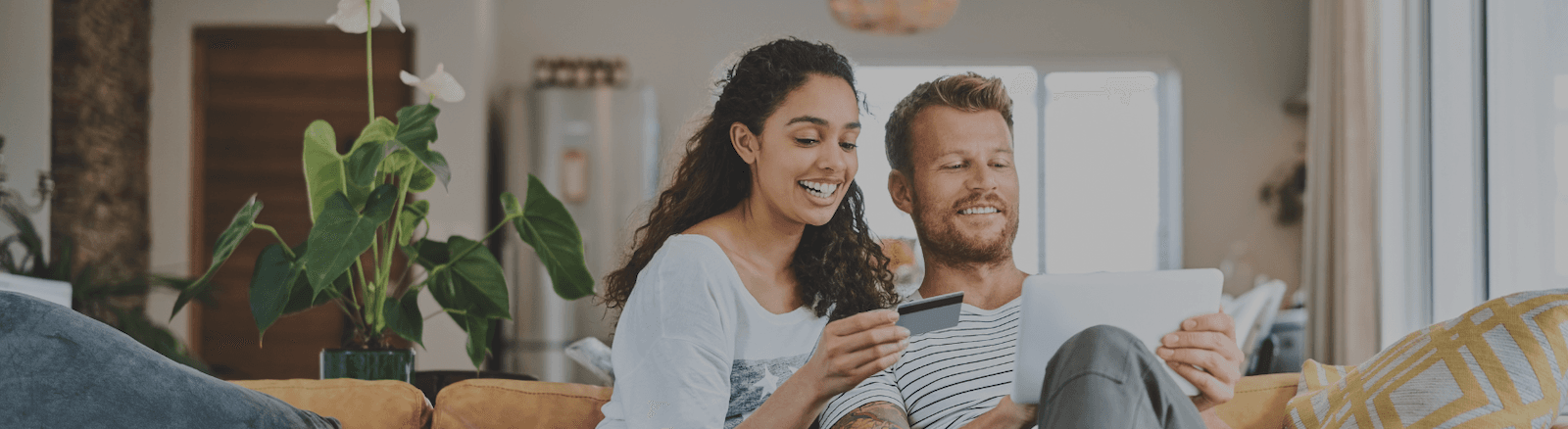 Smiling couple on couch shopping online with a tablet and credit card in a bright living room.