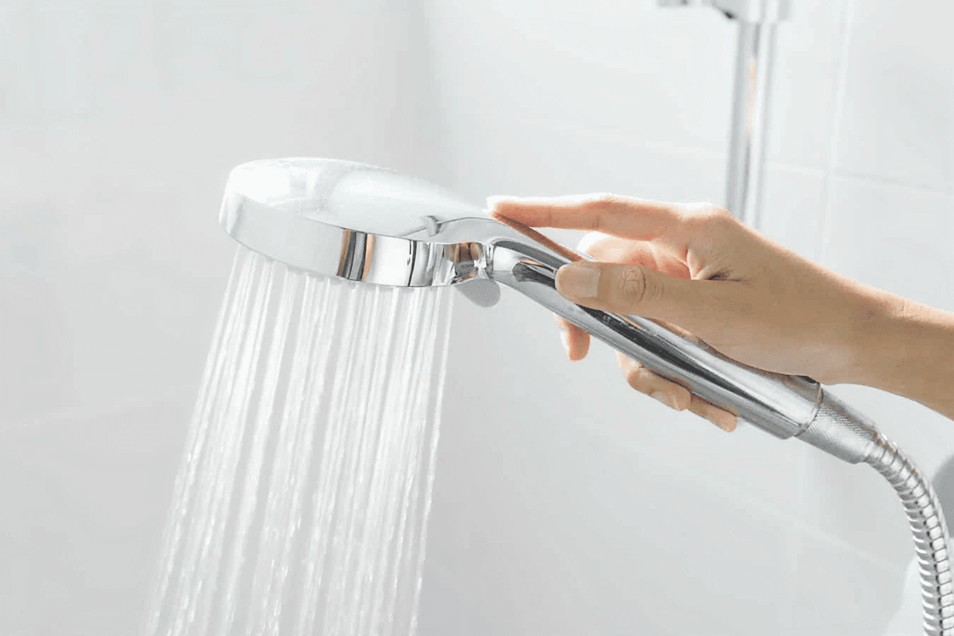 Hand holding a shower head with water flowing in a bright, tiled bathroom.