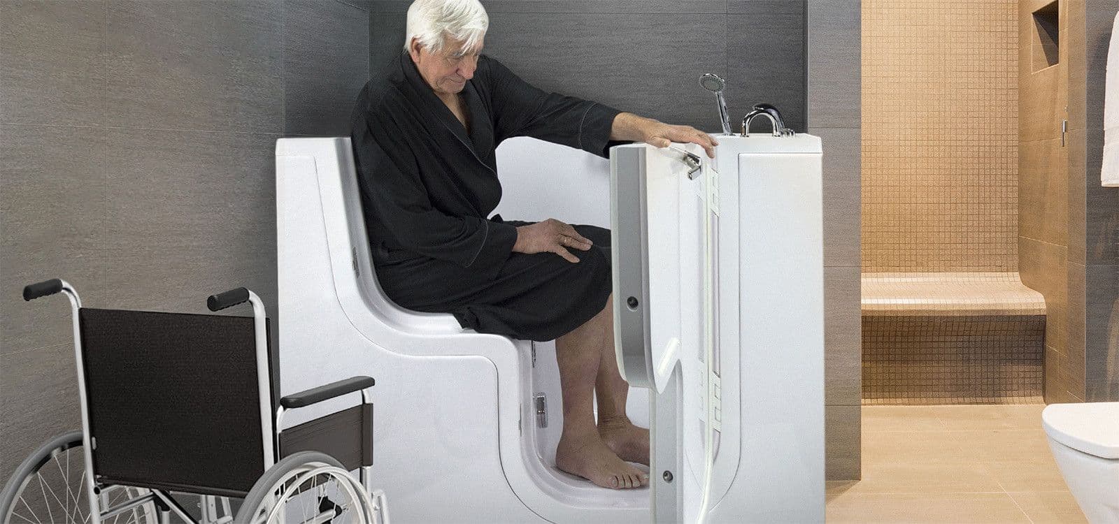 Older man in robe sits in a walk-in bathtub next to a wheelchair in a modern bathroom.
