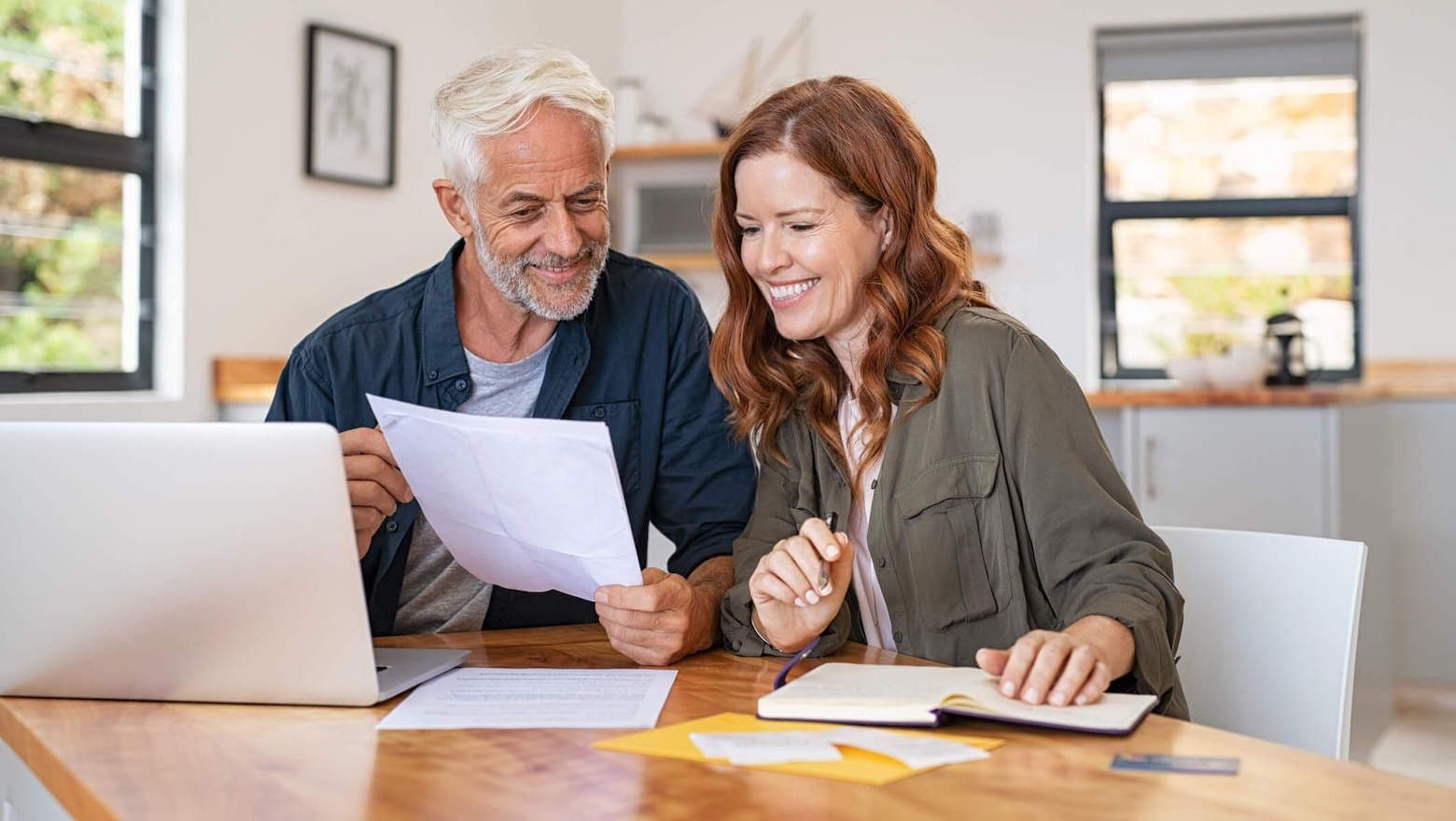 Smiling older couple at a kitchen table reviewing documents, with a laptop and notepad.