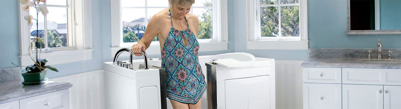 Older woman in a patterned dress steps into a walk-in bathtub in a bright, airy bathroom.