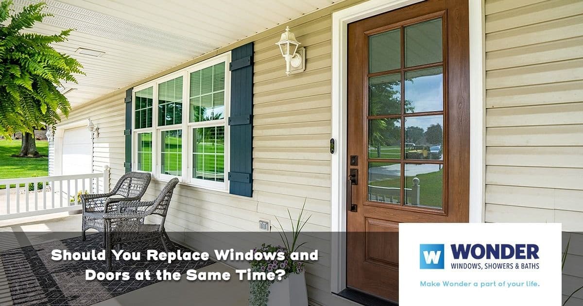 Charming porch with white siding, large windows, and a wooden door. A wicker chair and a doormat add a welcoming feel. Text reads: "Should You Replace Windows and Doors at the Same Time?"