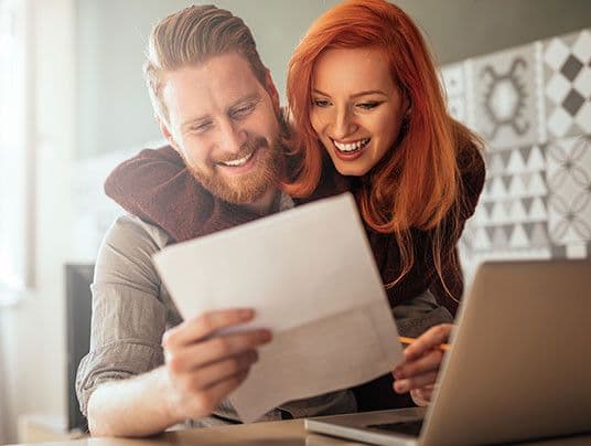 A happy couple is seated at a table, smiling as they read a document together. A laptop is open beside them.