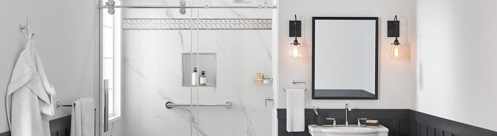 Modern bathroom with white marble shower, glass doors, and black trim. A sink with dual faucets is below an illuminated mirror. Towels and a robe hang nearby.
