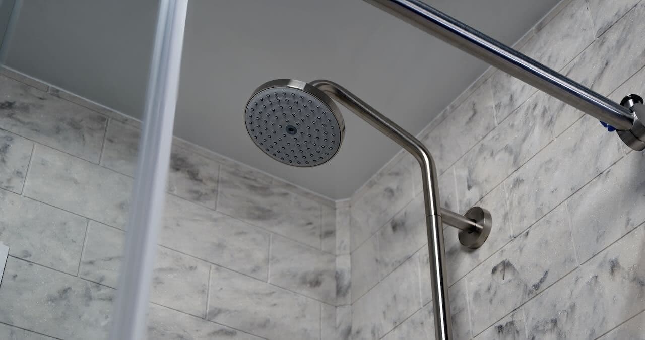 Close-up of a modern, round showerhead in a tiled bathroom with a marble-patterned wall.