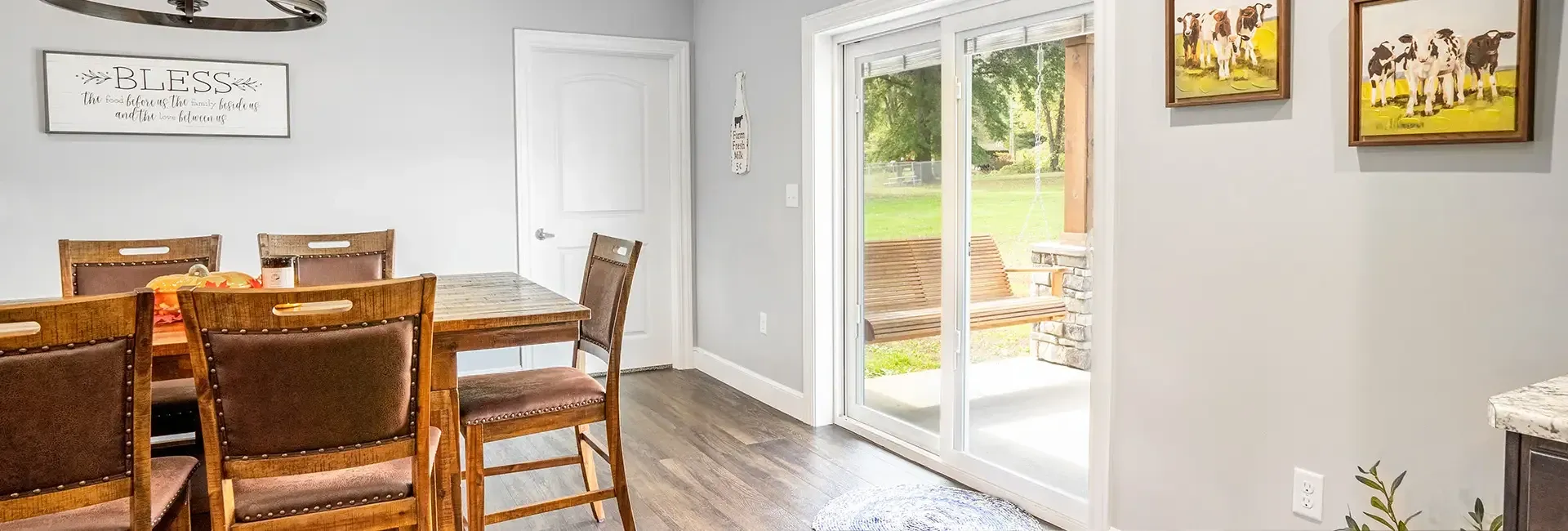 Bright, light gray dining area with wooden table and chairs on wooden flooring. A sliding glass door opens to a green yard.