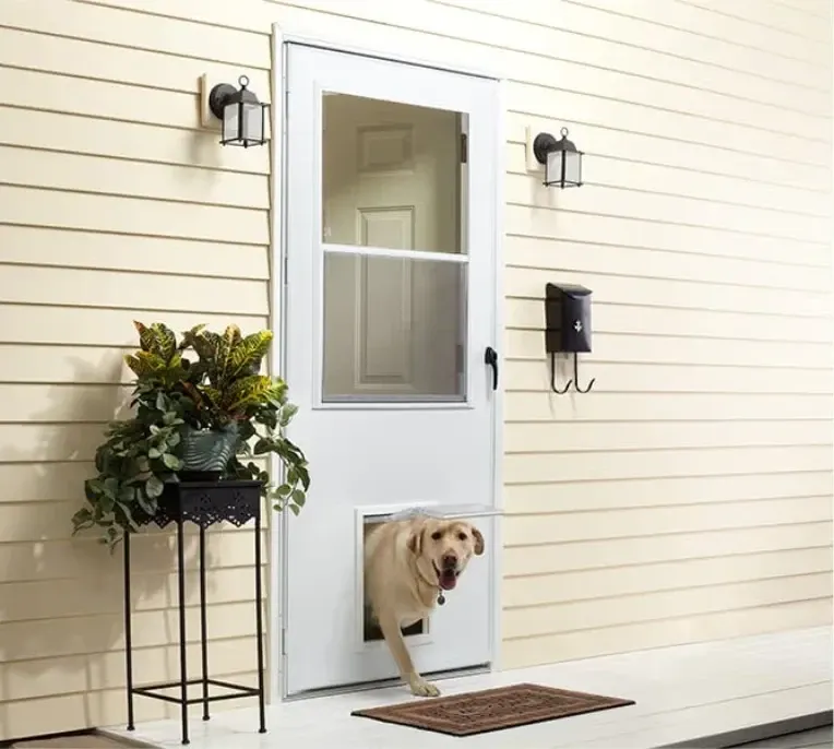 A dog happily exits through a white storm door's pet flap on a light yellow house.