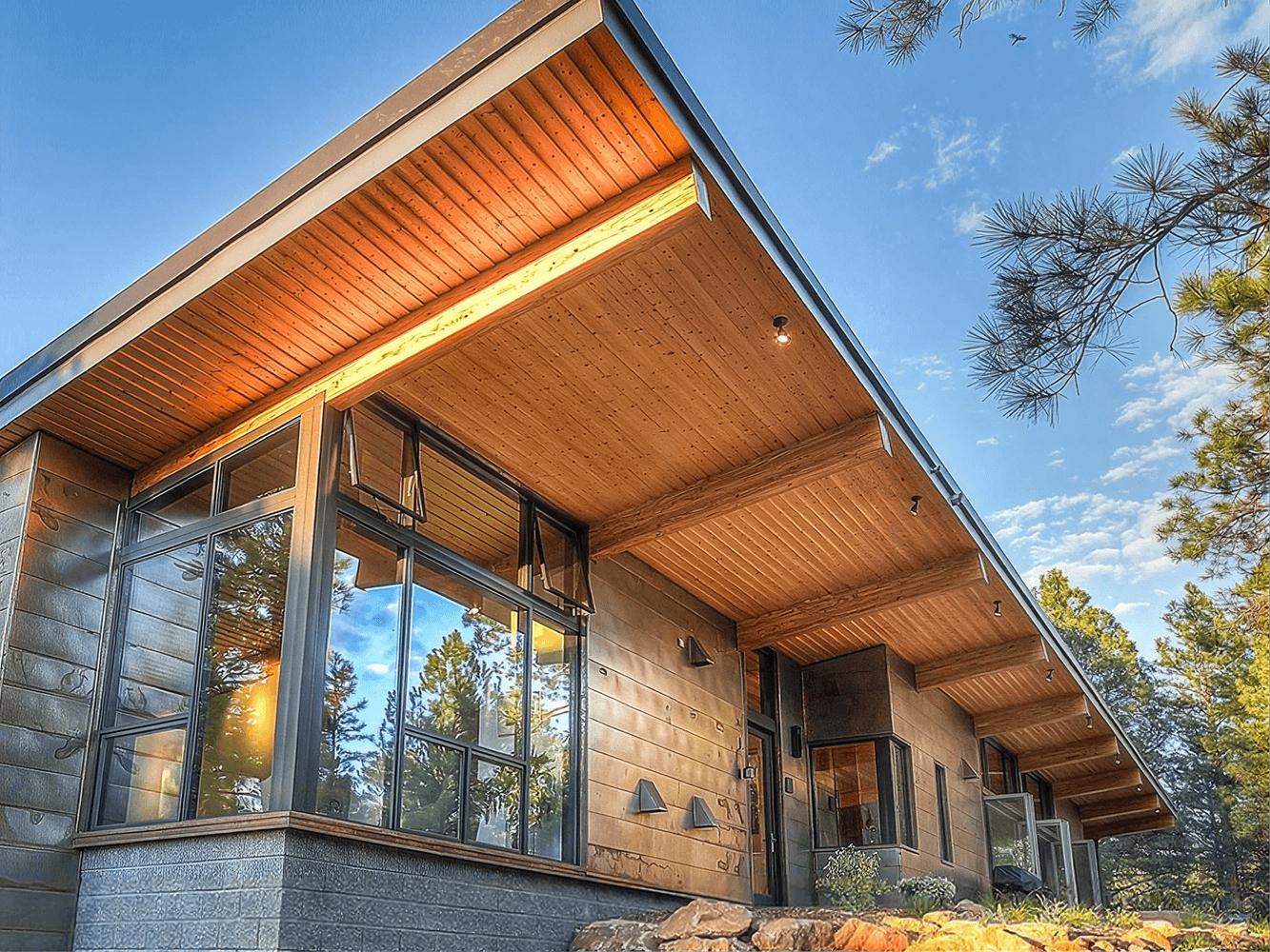 Modern residential home with copper-toned wood ceiling overhang, large black-framed glass doors and windows, stone foundation, surrounded by pine trees and blue sky.