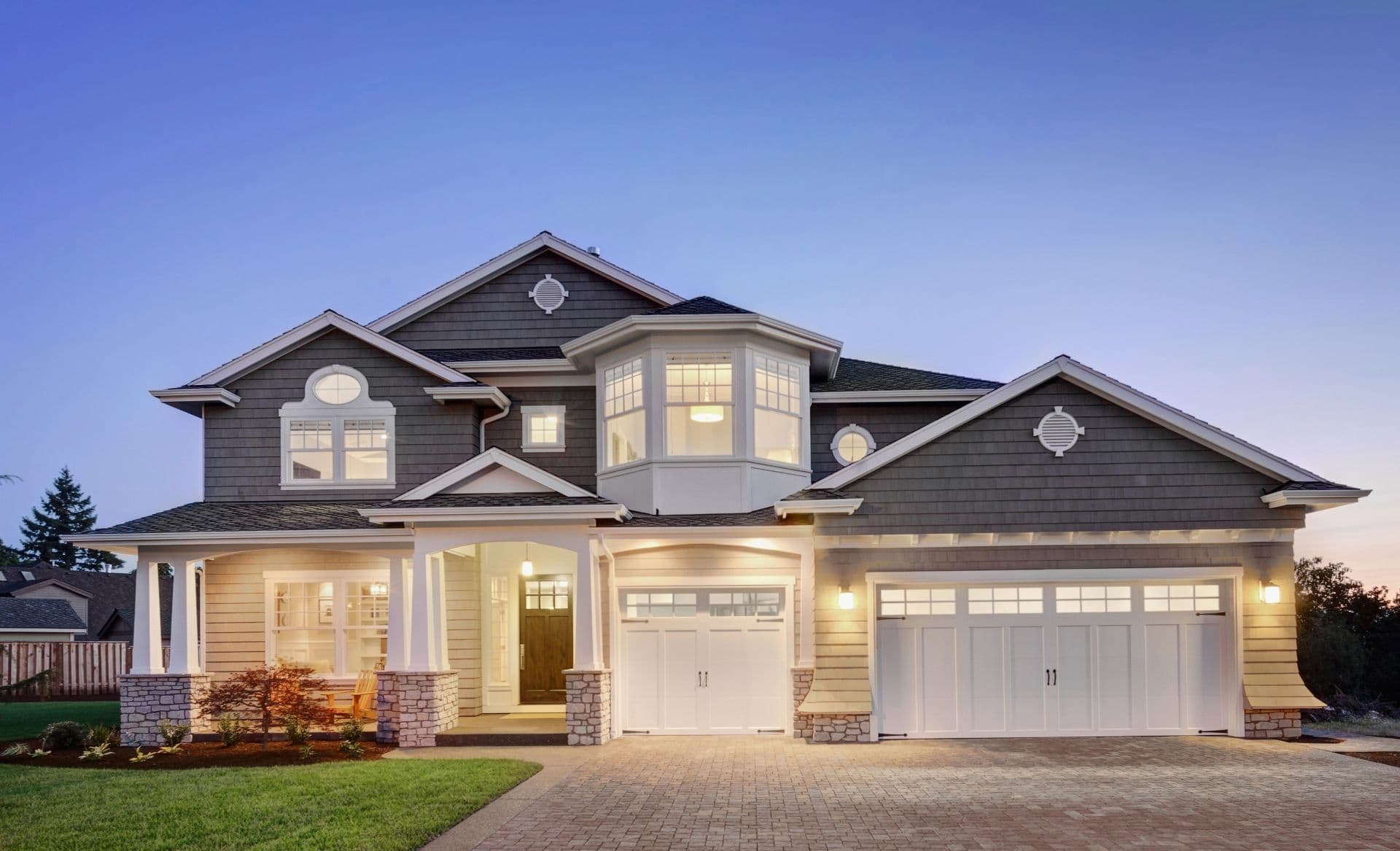 Modern two-story home with dark gray and tan exterior, white trim, stone accents, covered porch, and three-car garage lit at dusk.