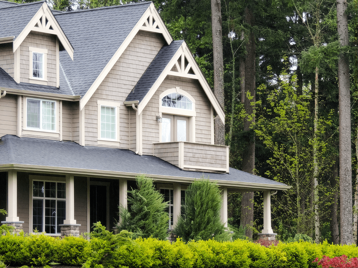 Two-story suburban home with taupe siding, gabled roof, front porch with columns, and surrounded by mature trees and green landscaping.