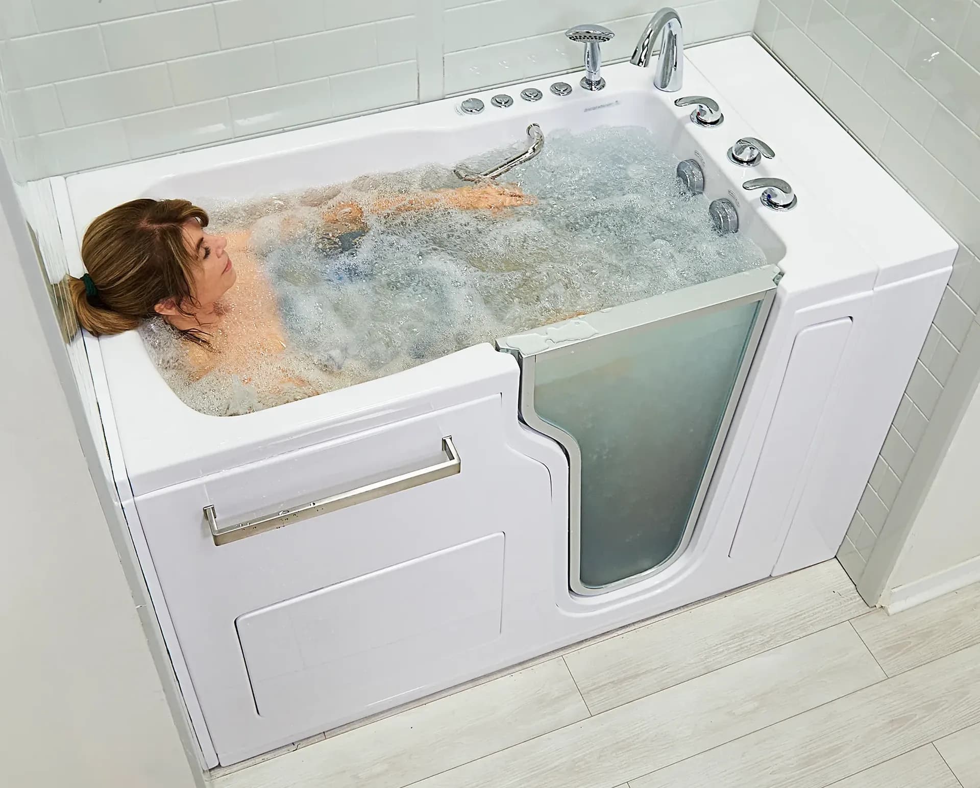 A woman relaxes in a partially filled walk-in bathtub with a glass door, surrounded by bubbling water.