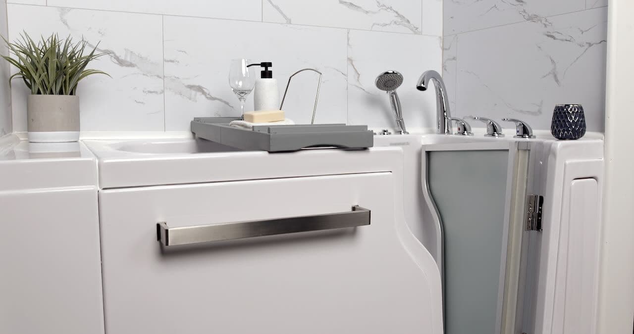 A modern, accessible bathtub with a safety door and stainless steel handle. It features a tray with soap, a plant, a glass, and a black soap dispenser, set against a white marble wall.