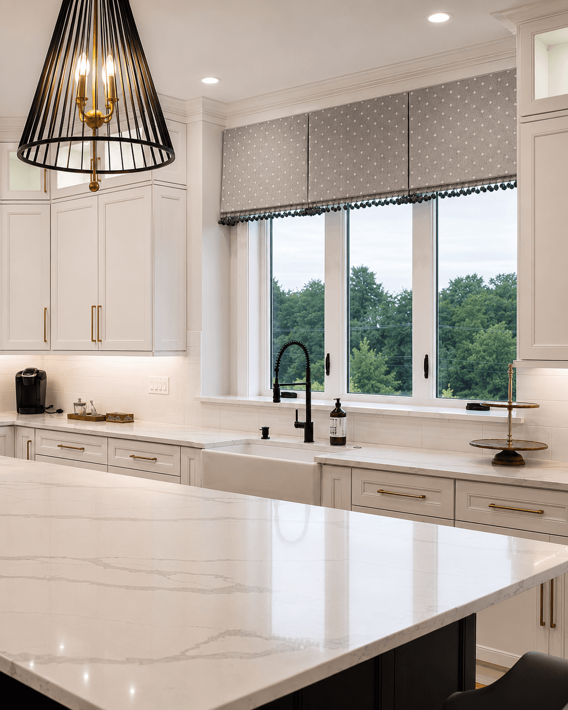 Modern kitchen with white cabinetry, marble countertops, black cone pendant light, and large casement windows with patterned roman shades overlooking trees.