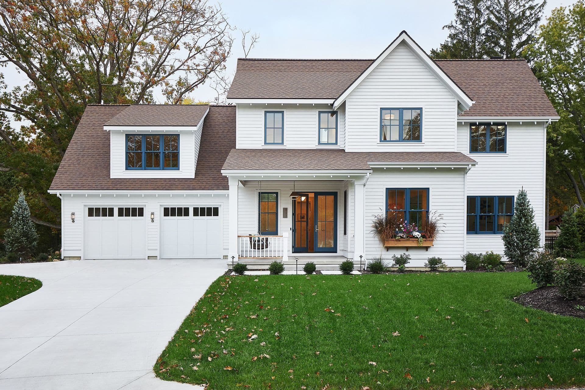 A modern two-story white house with a brown roof, blue trim windows, and a welcoming front porch. Set against a green lawn and surrounded by trees.