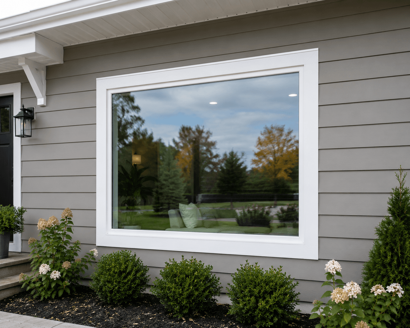 Large picture window with white frame on gray house exterior, showing view of landscaped backyard with trees and lawn, surrounded by flowering shrubs and plants.