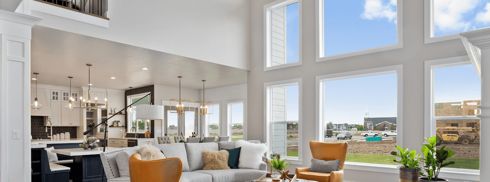 Open-concept modern home interior with two-story vaulted ceilings, gray sectional sofa with leather accent chairs, pendant lighting, and expansive picture windows overlooking rural landscape with construction equipment.