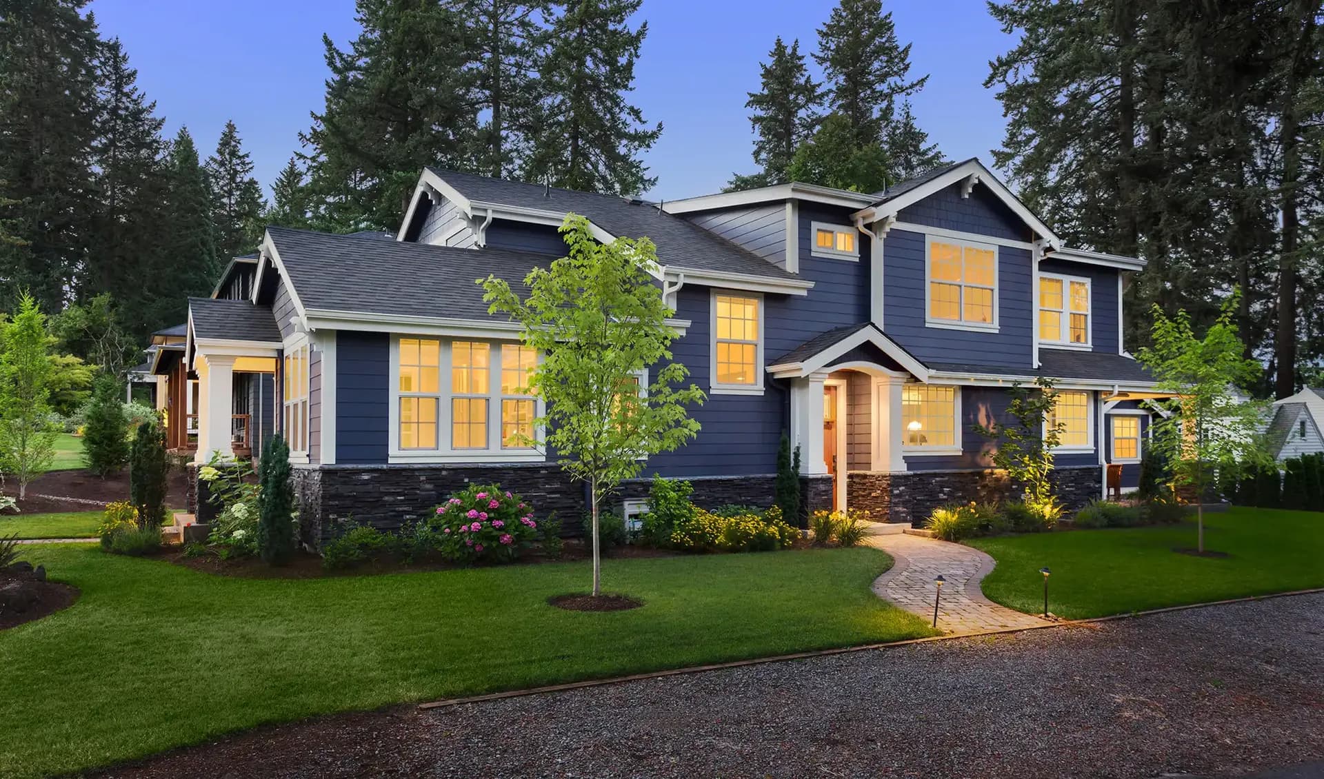 A charming blue two-story house with large windows warmly lit from inside, surrounded by lush greenery and tall trees.