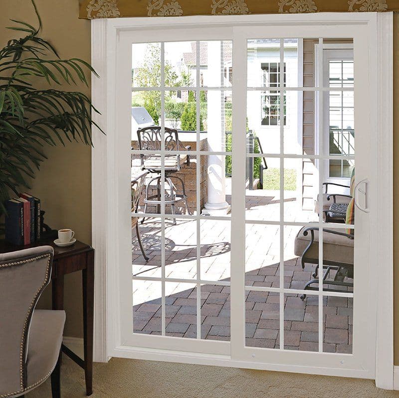 A white sliding glass door opens to a sunlit patio with brick flooring, outdoor furniture, and a barbecue grill, viewed from a cozy room with a potted plant.