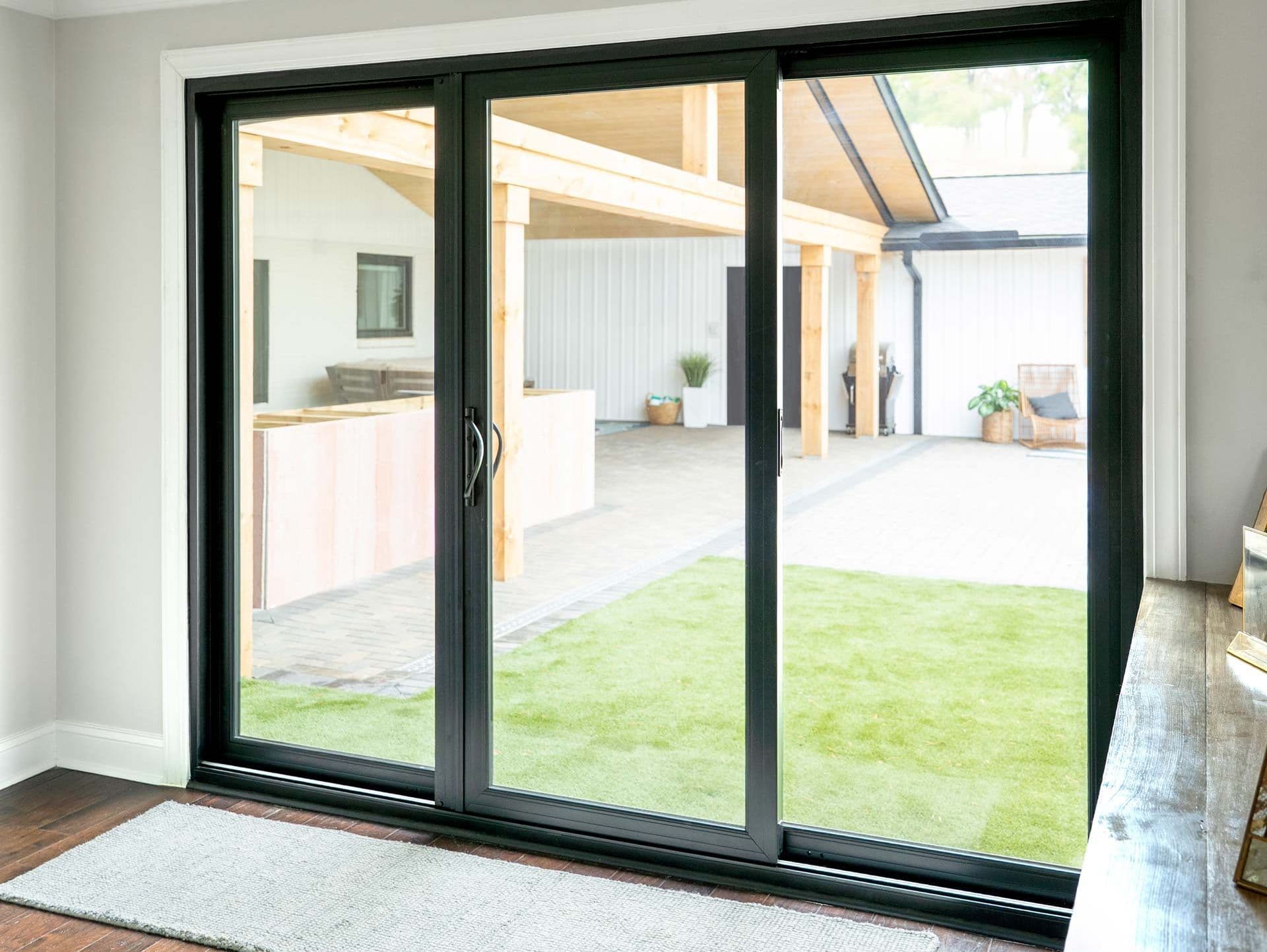 Sliding glass door with black frame opens to a bright modern patio. The outdoor area includes wooden beams and potted plants on a green turf.